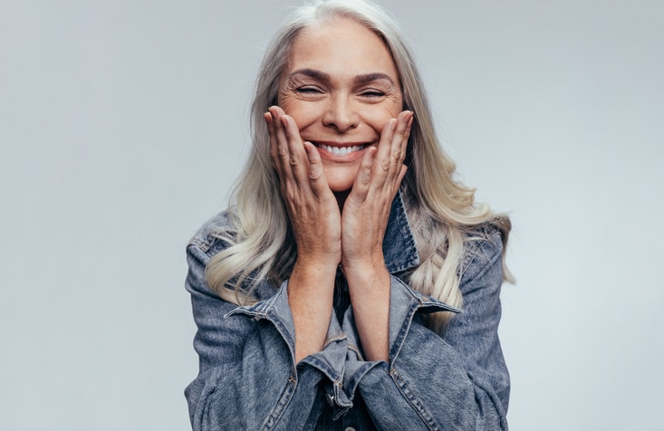 Dental Implants 3 Cheerful senior woman with hands on her face against grey background. Happy mature woman in casual denim shirt to help illustrate esthetic dentistry vs cosmetic dentistry