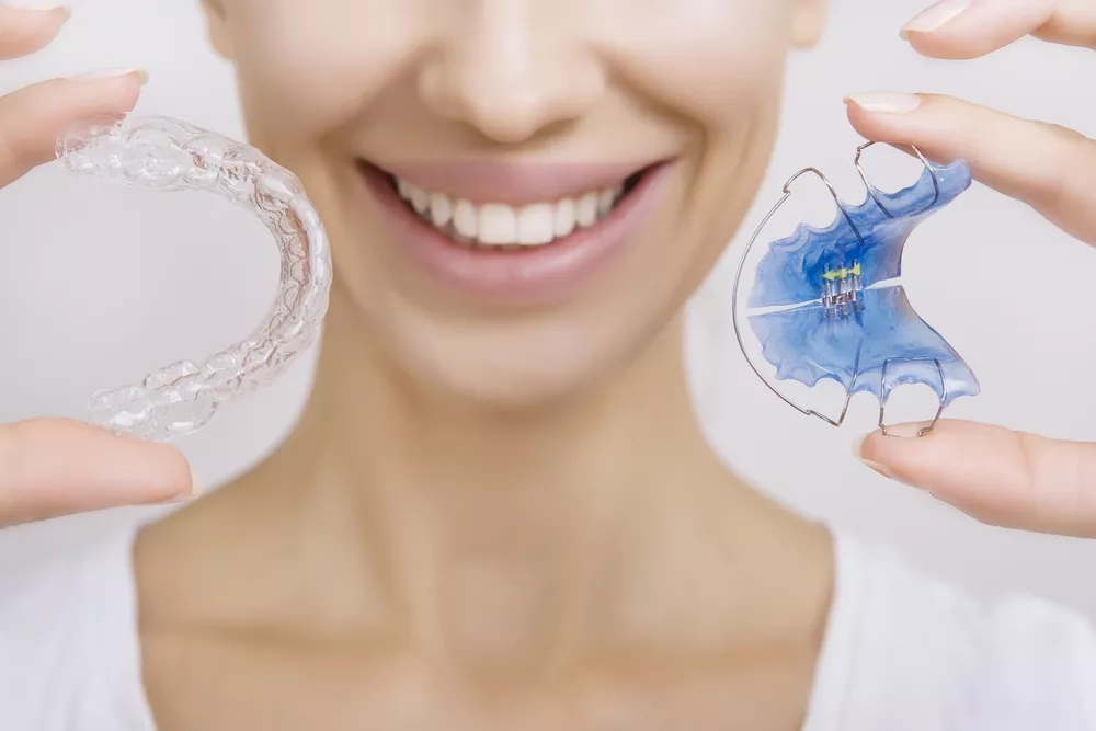 a smiling woman holding clear Invisalign braces in one hand and metal braces in the other